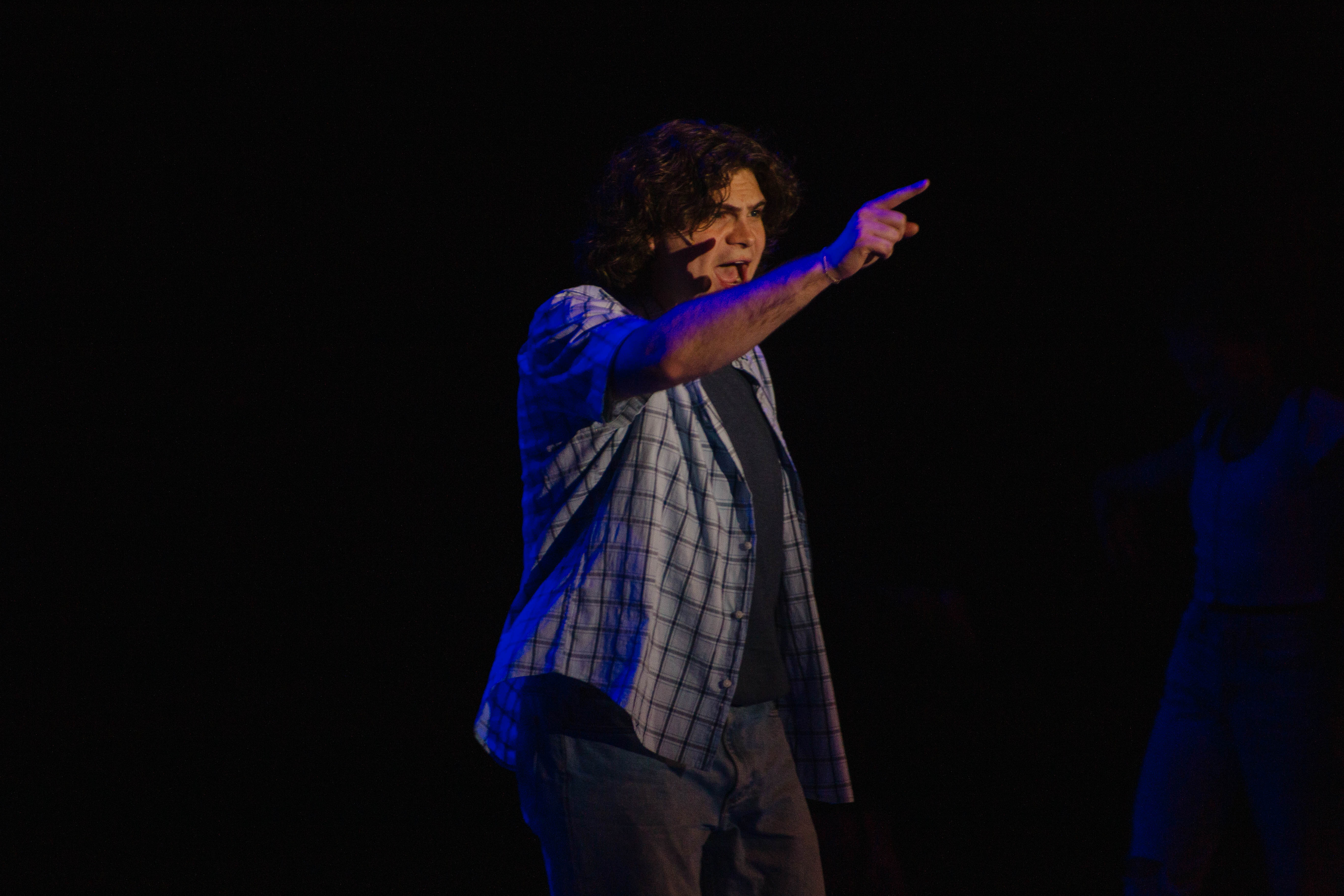Pointing across the stage in a blue plaid shirt